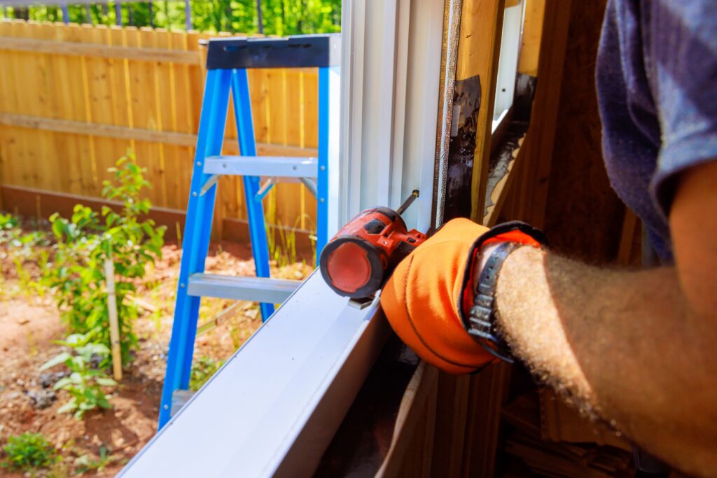 A contractor wearing orange work gloves uses a power drill to install a white door or window frame, with a blue step ladder visible in the backyard behind them.