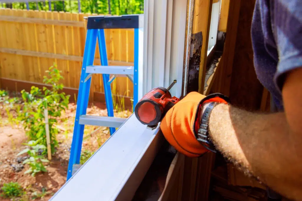 A contractor wearing orange work gloves uses a power drill to install a white door or window frame, with a blue step ladder visible in the backyard behind them.